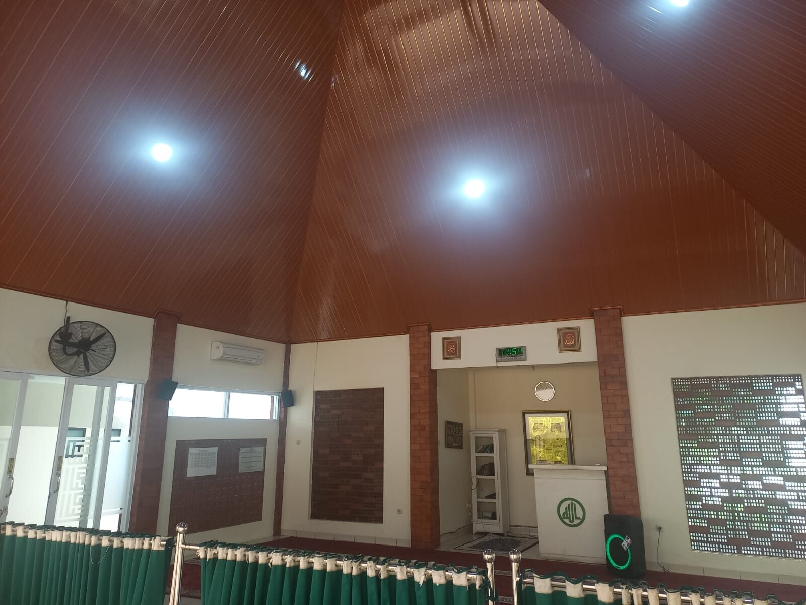 Ruang sholat dengan AC, kipas dinding, dan tirai hijau